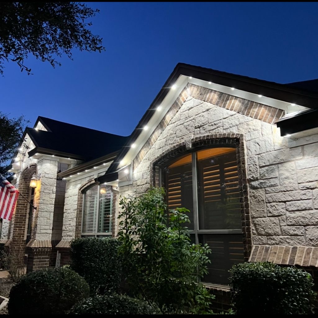 Stone-front home at dusk with cool white permanent lights along the roofline